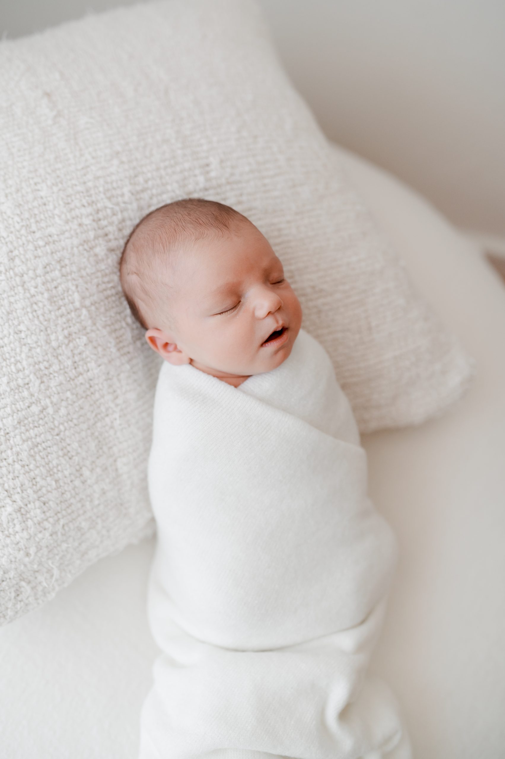 tiny newborn baby is wrapped up in a white swaddle and laid on a soft white pillow, she sleeps soundly with her mouth just slightly open. Beautiful creamy light floats down onto her cheek.