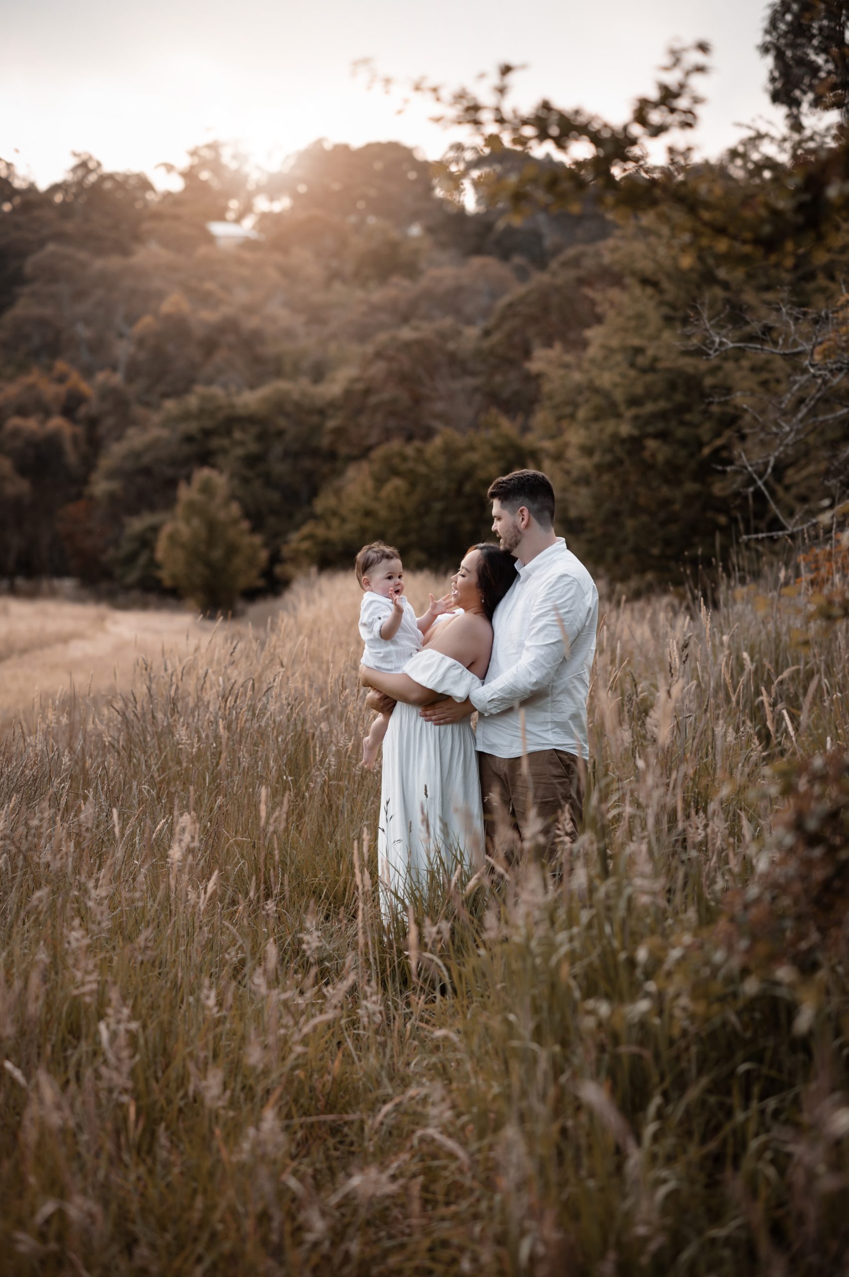 baby Oliver and his family are photographed at our Forest & Field location in Blackwood, South Australia. The evening is warm and the grass is long and wavy, the light from the sunset glows behind the family and falls softly on the field in front of them.